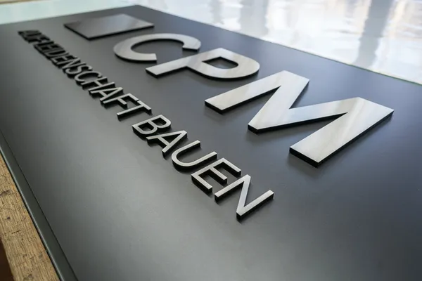 Ein Firmenlogo verklebt auf einer schwarzen Hartfaserplatte. Das Firmenlogo ist aus 8mm schwarzen Acryl gefertigt und zusätzlich mit 0.8mm gebürstetem Edelstahl beklebt.