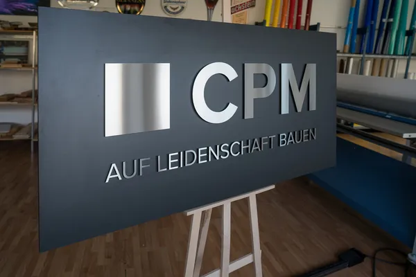 Ein Firmenlogo verklebt auf einer schwarzen Hartfaserplatte. Das Firmenlogo ist aus 8mm schwarzen Acryl gefertigt und zusätzlich mit 0.8mm gebürstetem Edelstahl beklebt.